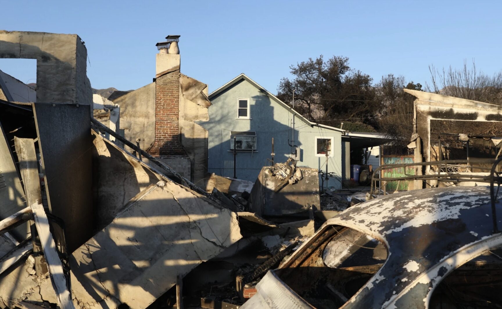 A scene showing the aftermath of a fire with a partially destroyed house and charred remains of cars. Burnt walls and a chimney are visible, while a blue intact house stands in the background under a clear sky.